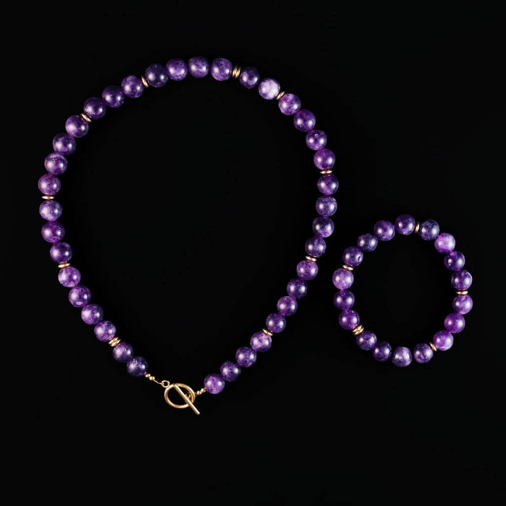 A matching beaded necklace and bracelet set in purple with small gold spacer beads and gold-toned hardware, displayed on a solid black background.
