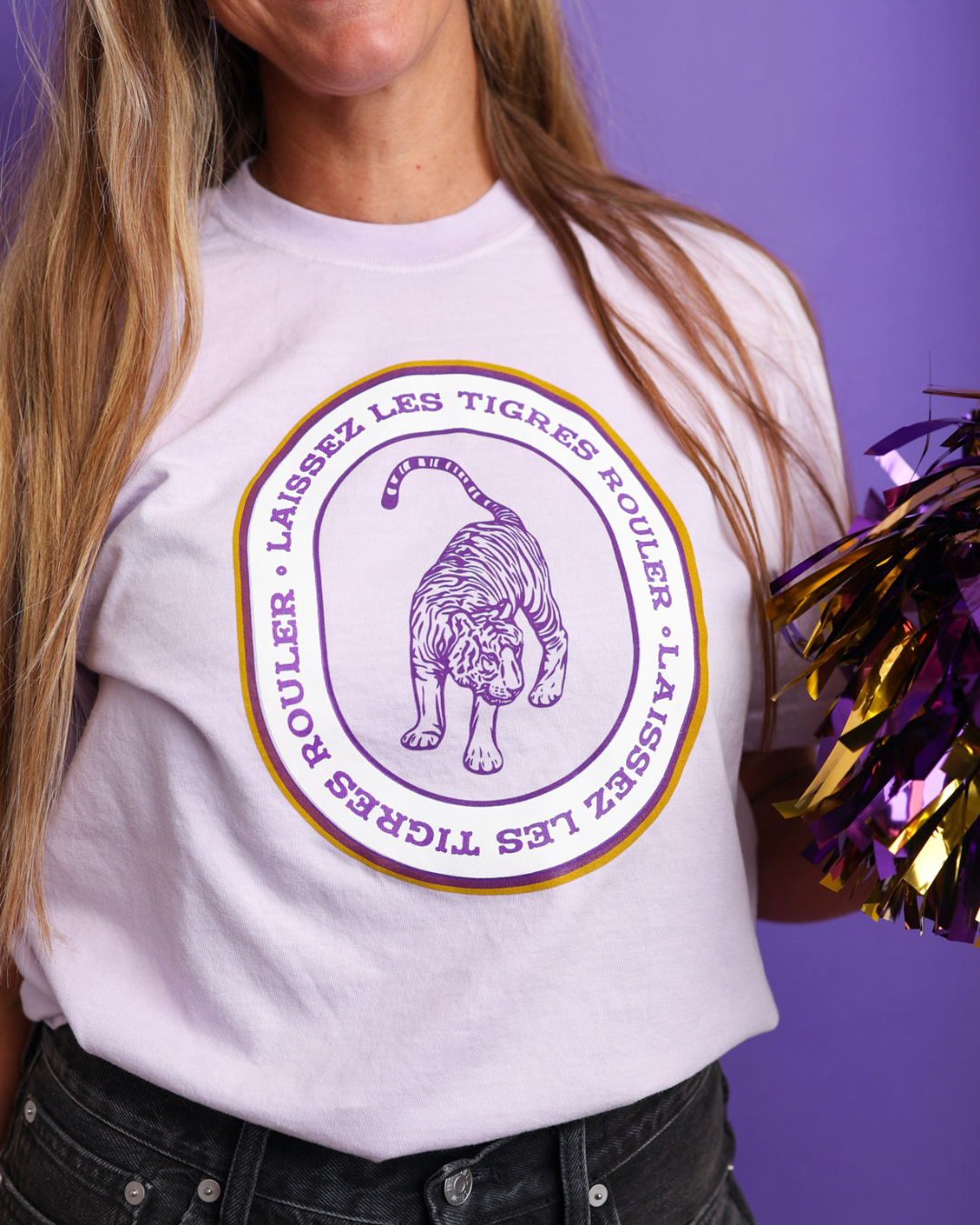 A person wearing a Lavender  graphic T-shirt with a purple and gold tiger illustration and French text, paired with dark jeans against a purple background.