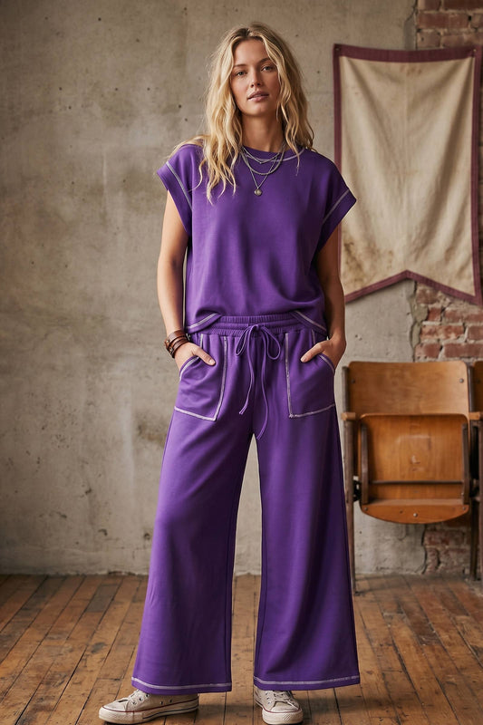 A woman modeling a matching purple lounge set consisting of a short-sleeve top and wide-leg drawstring pants with white piping, standing in a rustic room with a concrete wall and wooden floor.