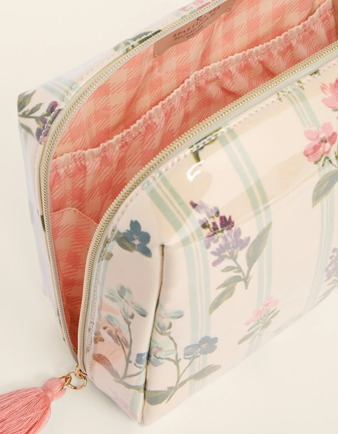 A close-up of a cosmetic pouch with a floral and striped print, featuring a pink gingham lining, a gold zipper, and a pink tassel.