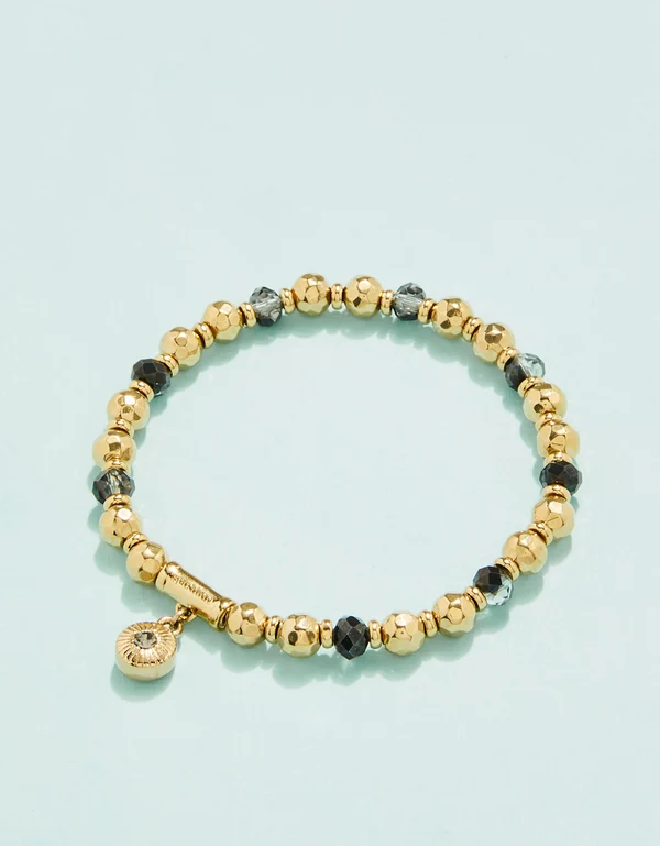 A gold beaded stretch bracelet with faceted black and gray beads, featuring a small round gold charm with a clear stone, displayed on a light blue background.