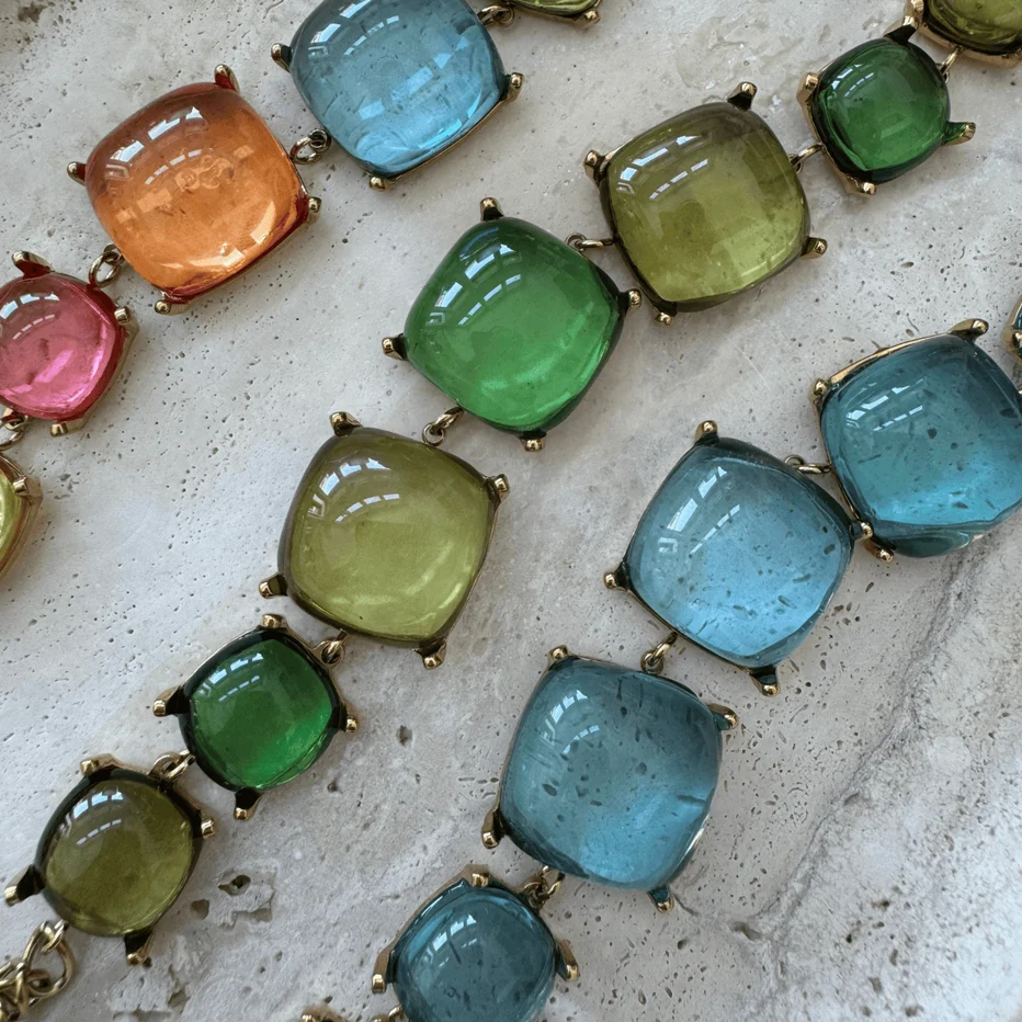 A close-up of a chunky beaded necklace featuring large, rounded square cabochon gemstones in multiple colors including blue, green, orange, and pink, set in gold-toned metal on a textured light stone background.
