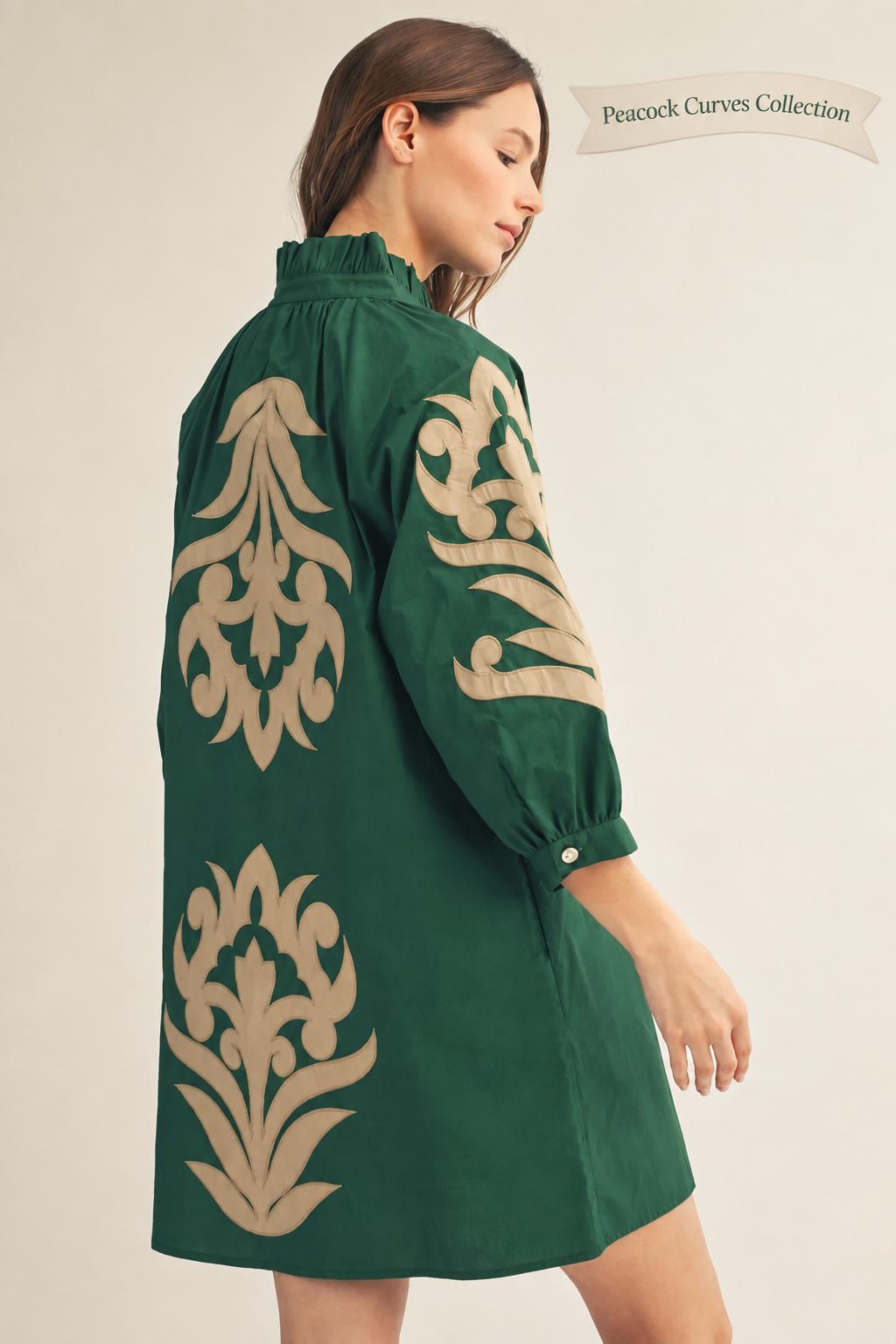 A woman wearing a dark green shift dress with large beige damask-style embroidery on the back and sleeve, shown from a side-back angle against a plain light beige studio background.