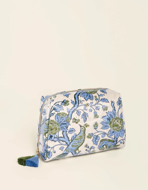 A small zippered cosmetic pouch with a blue and green floral and bird pattern on a cream background, featuring a blue and green tassel zipper pull, photographed on a light cream background.