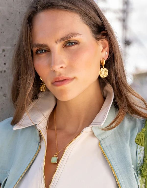 A close-up portrait of a woman wearing gold-toned drop earrings with circular medallion details, a gold necklace with a light green square pendant.