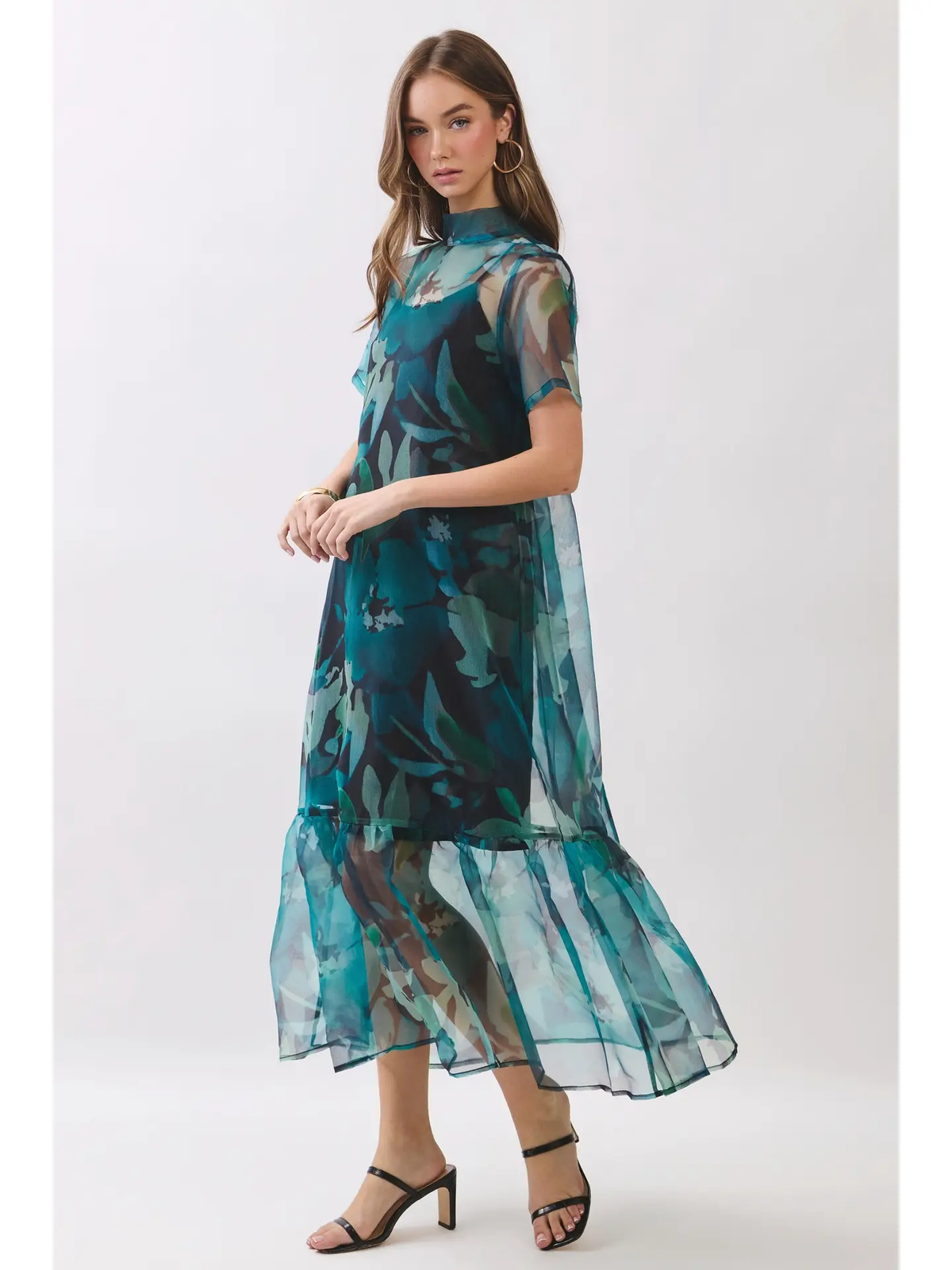 A woman modeling a sheer midi dress with a teal and green abstract floral print, featuring short sleeves, a high mock neck, and a tiered ruffled hem, paired with black strappy heels against a light gray studio background.