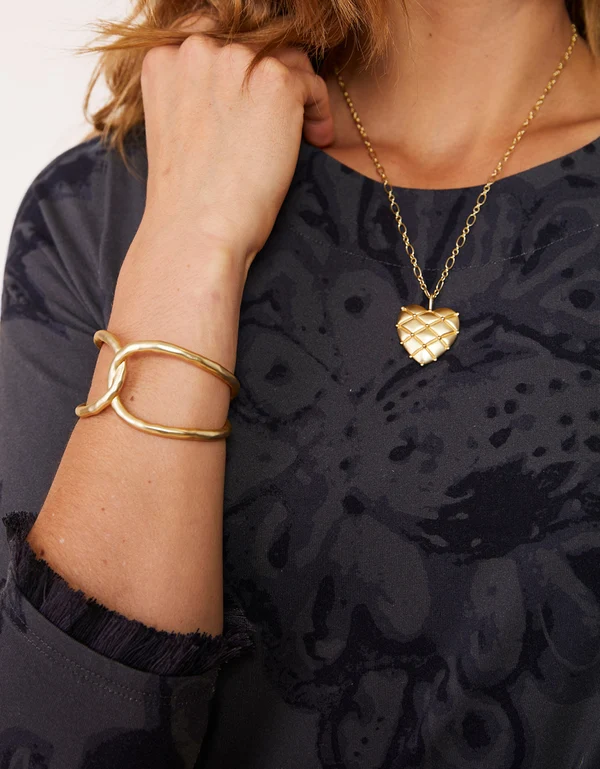 A close-up lifestyle shot of a model wearing a gold heart-shaped pendant necklace and a matching twisted gold cuff bracelet.