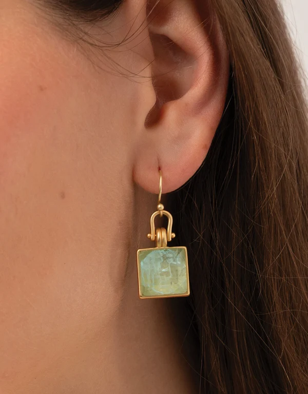 A close-up of a person's ear wearing a gold dangle earring with a square light green stone.