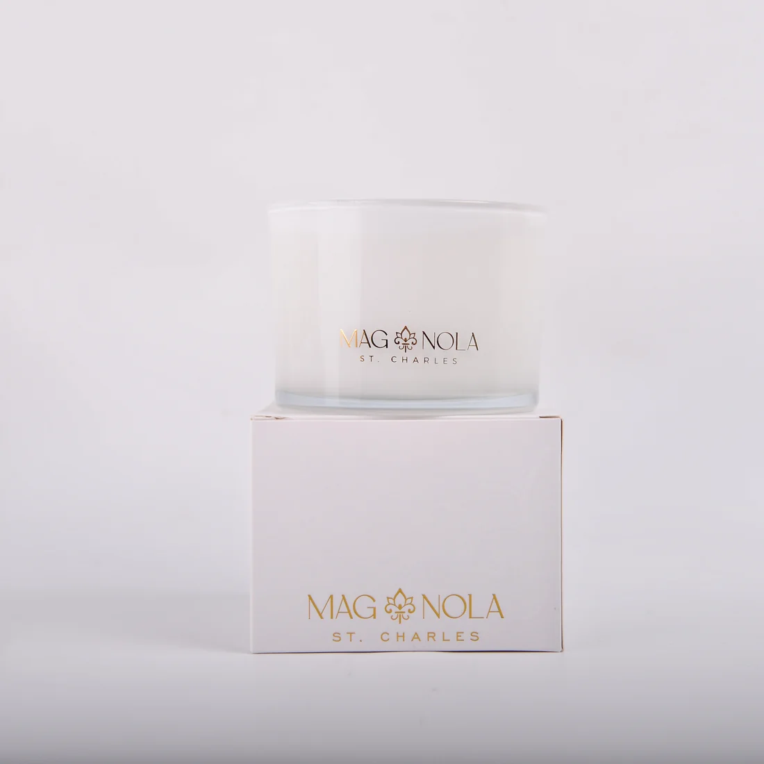A frosted 14 oz glass jar candle with a clear lid, featuring the brand name 'MAG NOLA ST CHARLES' printed in gold on the front, set against a clean white background sitting on top of its container. 