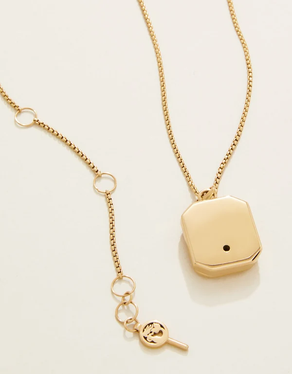 A gold necklace with a geometric pendant, shown alongside a matching chain extension featuring circular rings and a small key charm, on a clean off-white background.