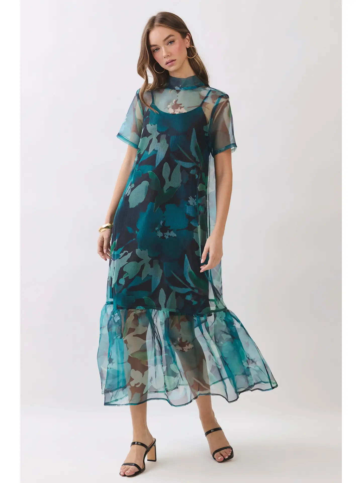 A woman modeling a sheer midi dress with a dark teal and green floral print, featuring short sheer sleeves, a high neck, and a ruffled hem, paired with black heeled sandals against a light gray studio background.