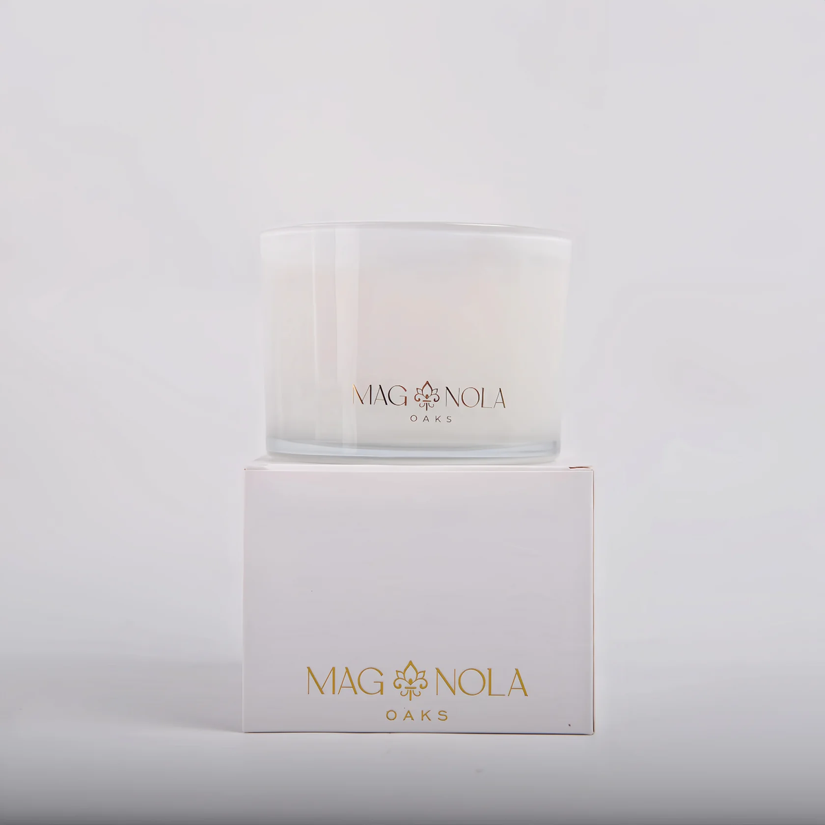 A clear glass jar candle with white wax sits on top of its white box, both printed with the brand name MAG NOLA OAKS in gold lettering, against a clean white background. 