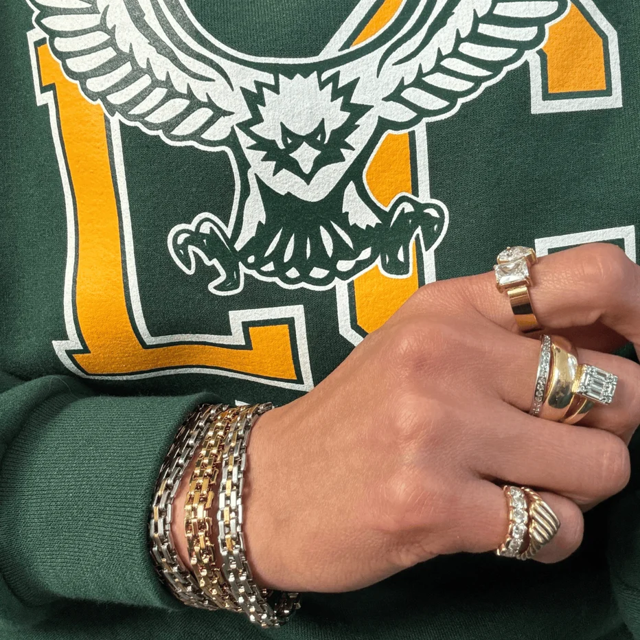 Hand with gold rings and bracelets on a green background with an eagle logo.