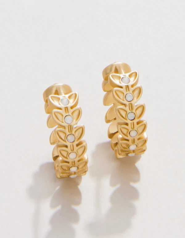 A pair of gold hoop earrings with a repeating leaf pattern and small white circular accents, displayed on a clean white background with soft shadows.
