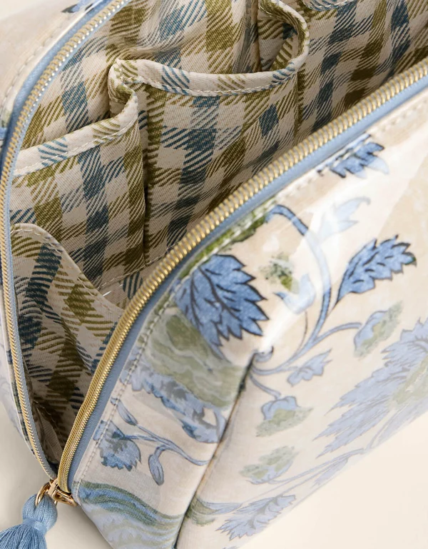 Close-up of a toiletry bag showing its cream-colored exterior with a blue botanical pattern and its matching patterned interior, featuring a gold zipper and a blue tassel pull.