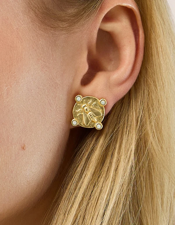 A close-up of a model's ear wearing a gold stud earring with a raised bee motif and small white pearl-like accents.