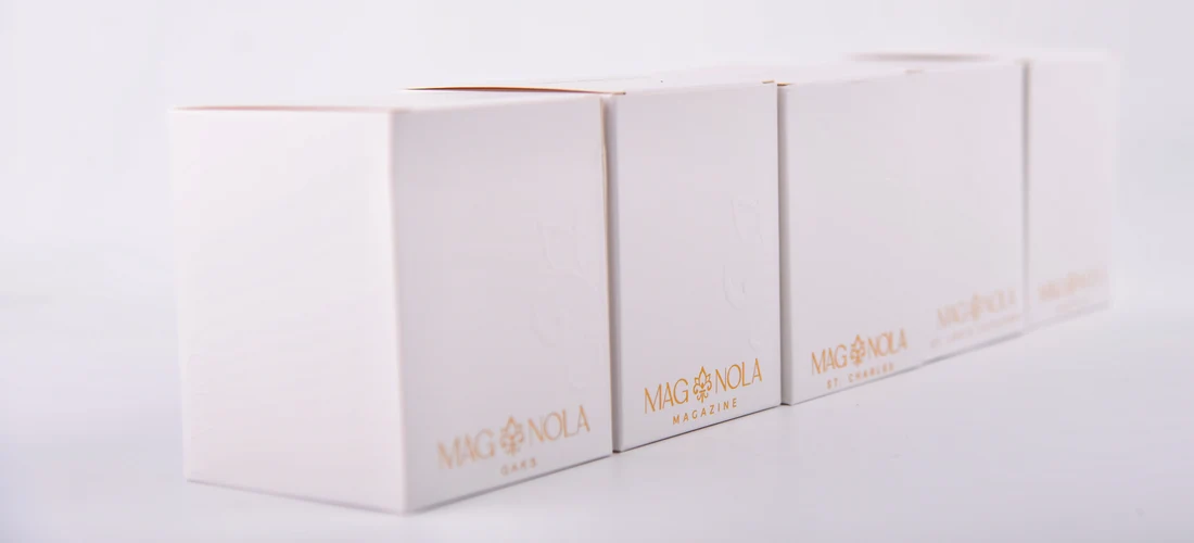 White rectangular 8.5 oz candle boxes arranged in a row on a white background, each printed with the brand name MAG NOLA in gold lettering.