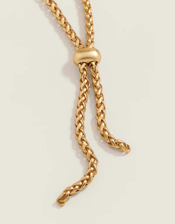 A close-up of a gold-toned chain necklace with a central knot and a smooth oval connector, photographed on a plain light beige background.