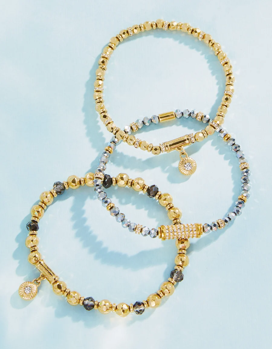 Three beaded stretch bracelets arranged in a circular pattern on a light blue background. Two bracelets are gold with dark gray faceted beads and a small round crystal charm, while the third is silver with clear crystal beads and a crystal-encrusted clasp.