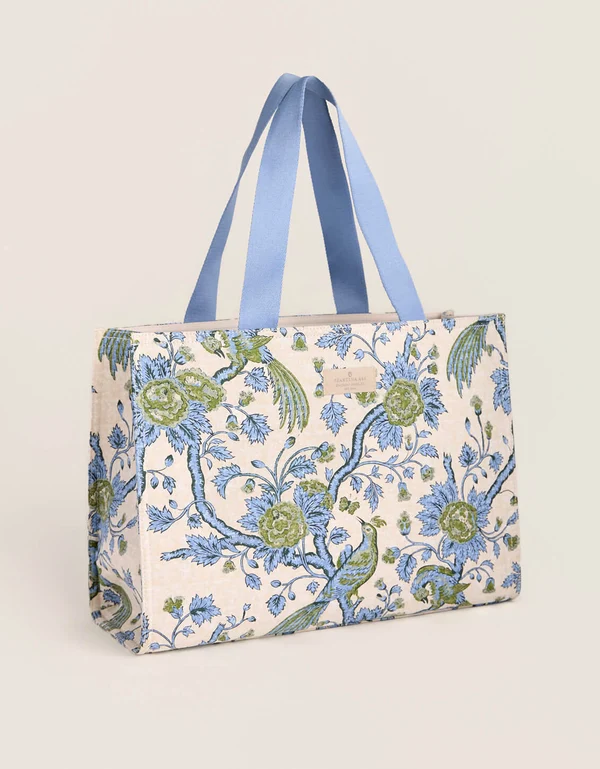 A tote bag featuring a blue and green floral tapestry pattern on a cream background, with light blue fabric shoulder straps, shown against a solid light beige background.