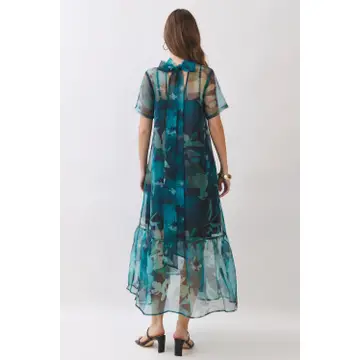 A woman viewed from the back wearing a sheer midi dress with a blue and green abstract print, paired with black heeled sandals, against a plain light gray background.