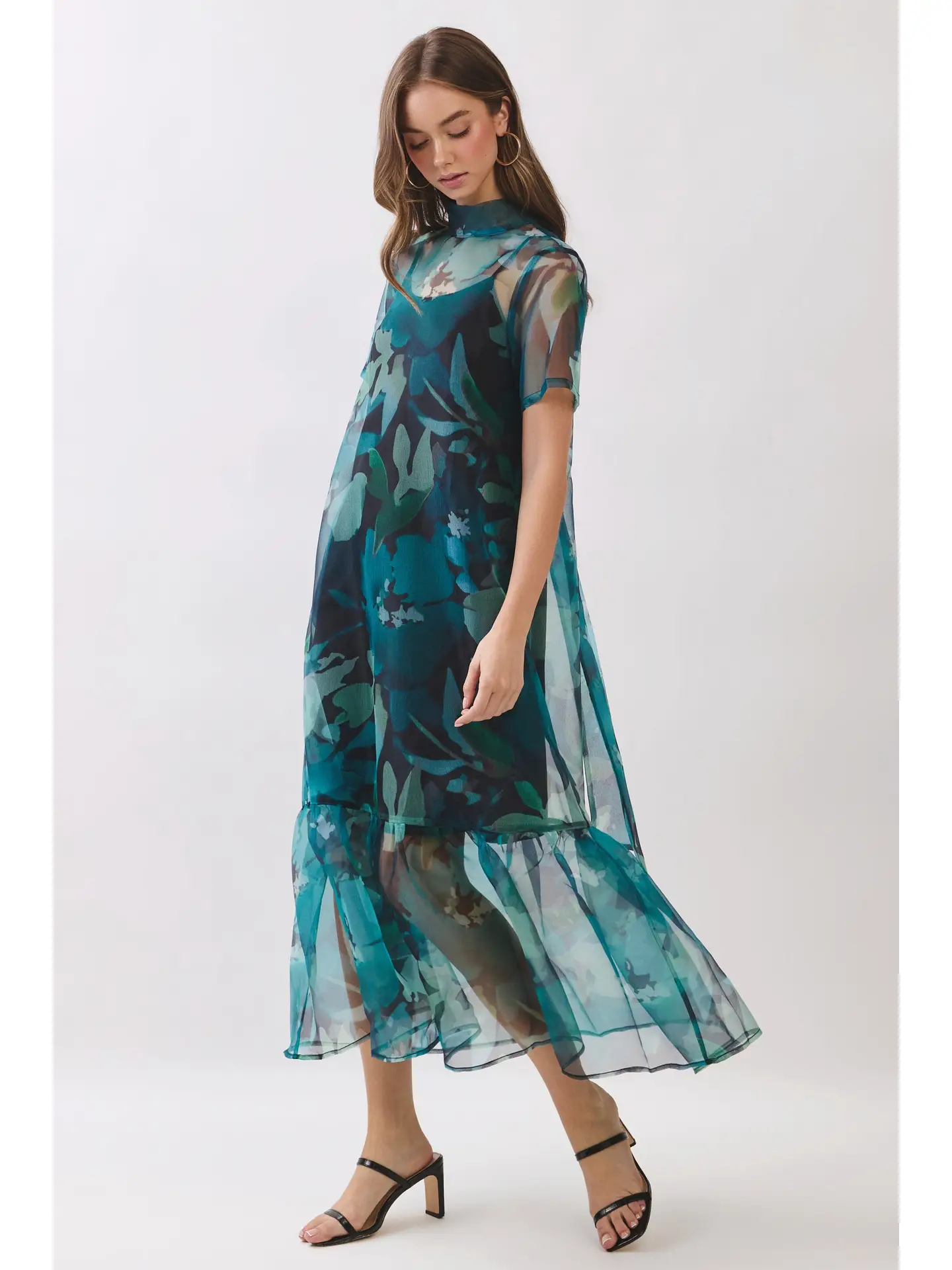 A woman modeling a sheer midi dress with a blue and green floral print, paired with black strappy high-heeled sandals, against a plain light gray studio background.