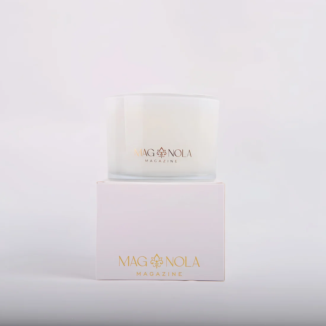 A frosted 14 oz glass jar candle with gold 'MAG NOLA MAGAZINE' branding sits centered on top of a pale pink square box, also branded in gold, against a clean white studio background.