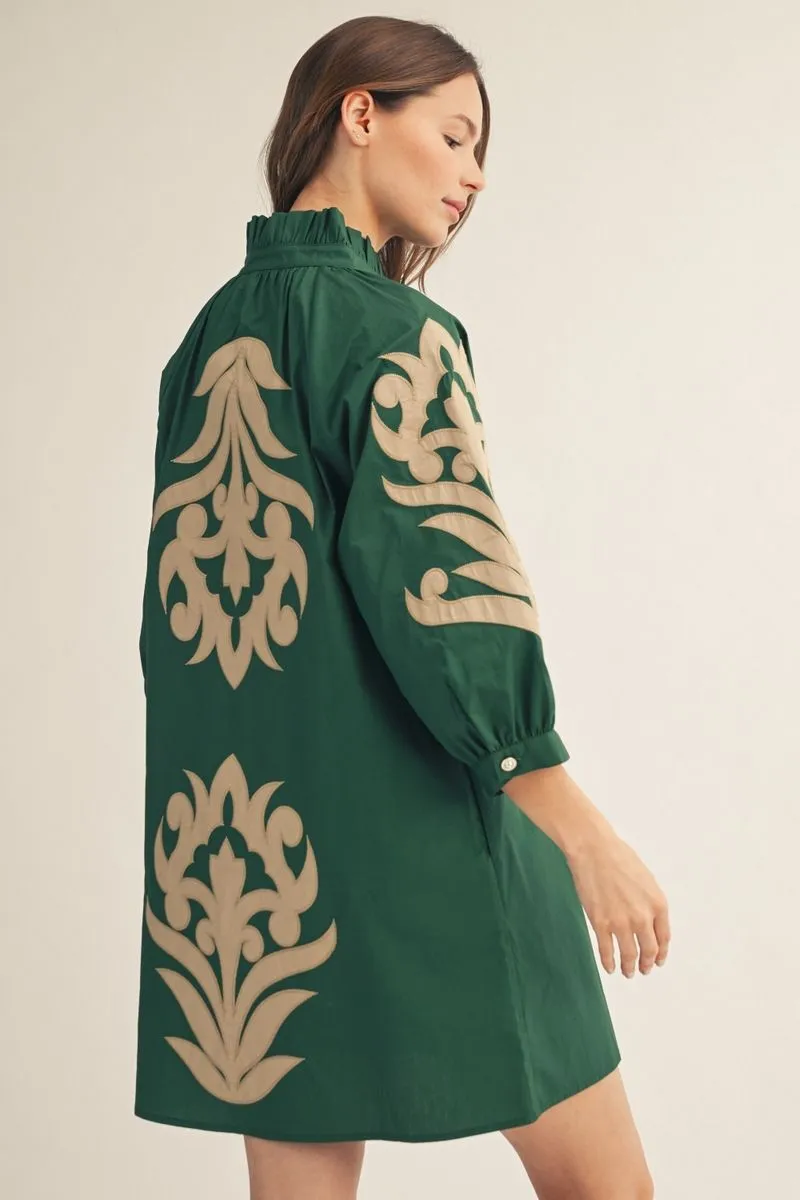 A woman wearing a dark green shift dress with large beige damask-style embroidery on the back and sleeve, shown from a side-back angle against a plain light beige studio background.
