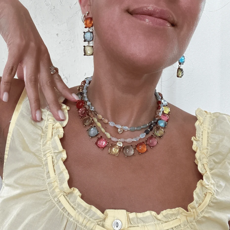 A close-up of a person wearing a multi-layered beaded necklace with colorful rectangular gemstones and matching drop earrings, paired with a pale yellow ruffled blouse.