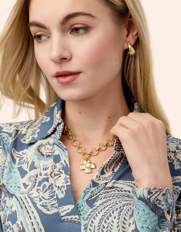 A close-up of a woman wearing a gold cross pendant necklace and a matching gold hoop earring, paired with a blue and white paisley patterned blouse.