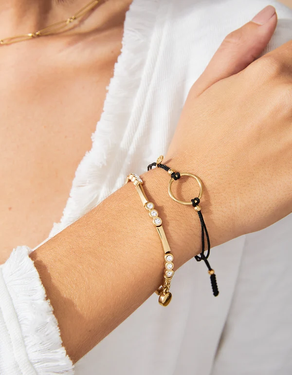 Close up of model wearing god bracelet featuring rhinestones and small gold dangle charm. 