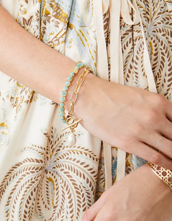 Close-up of a hand wearing a gold bracelet with turquoise beads and a pearl charm, against a patterned fabric background.