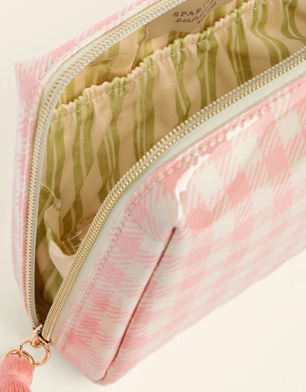 A close-up of a pink and white patterned cosmetic pouch with a gold zipper, shown open to reveal a green and beige striped interior lining. A small pink tassel charm is attached to the zipper pull.