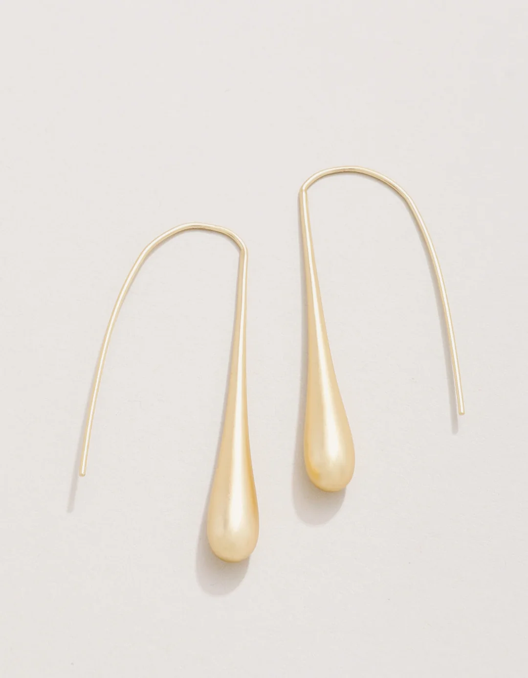 A pair of gold teardrop dangle earrings with long curved ear wires, displayed on a clean, light beige background.