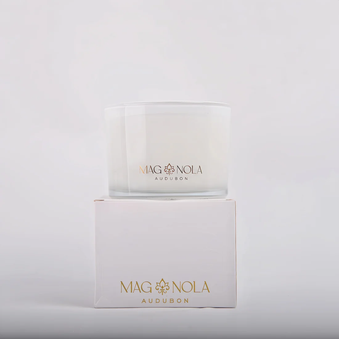 A frosted 14 oz glass jar candle with gold lettering reading 'MAG NOLA AUDUBON' sits on top of its white box, which also features the same gold branding, against a clean white background.