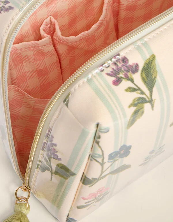 Close-up of an open zippered cosmetic pouch with a cream exterior printed with green leaves and purple flowers, a pale blue stripe, and a peach-pink patterned interior lining.