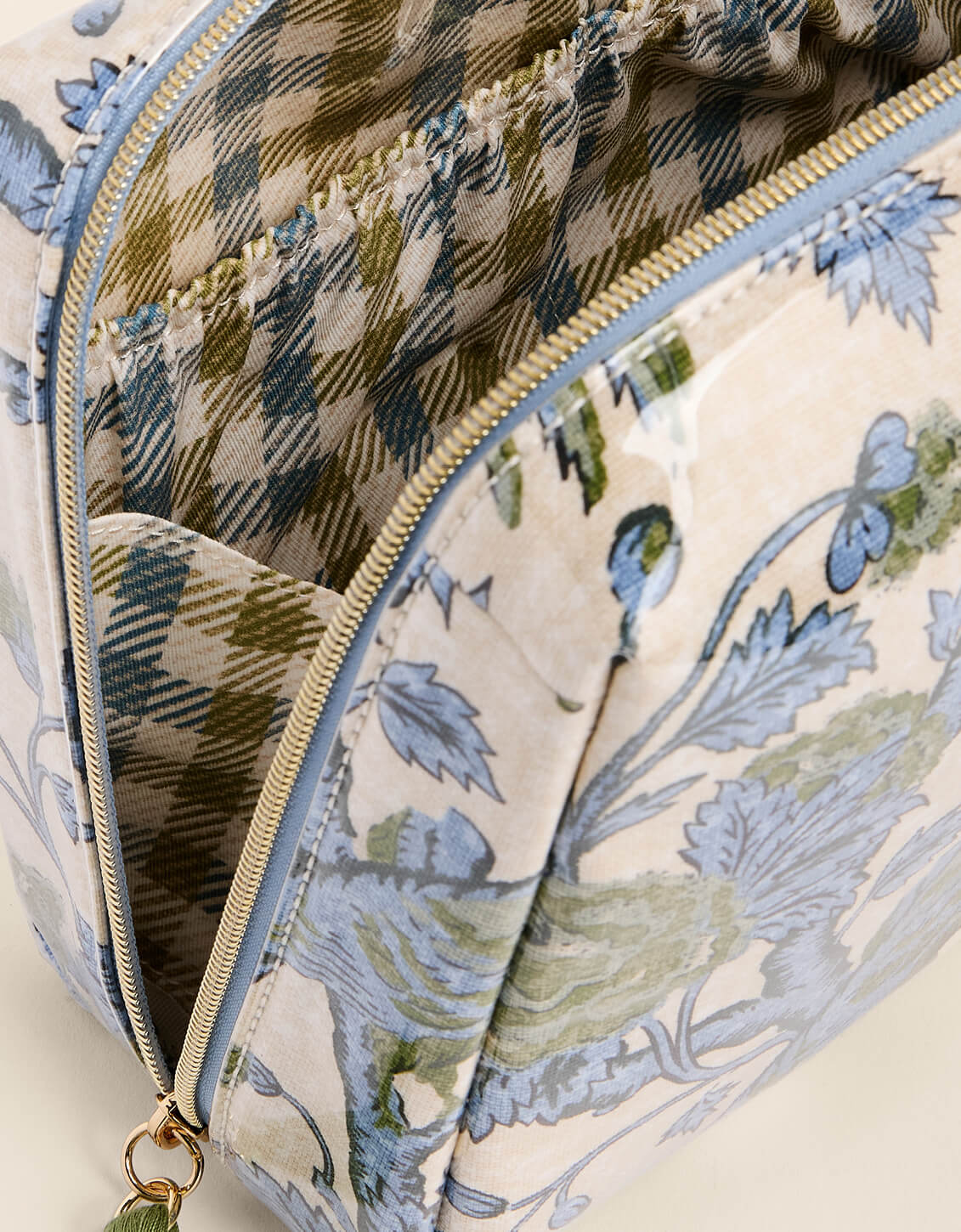 Close-up of a fabric zippered pouch or bag showing a cream-colored exterior with a blue and green botanical print, a blue and green plaid interior lining, and a gold-tone zipper.