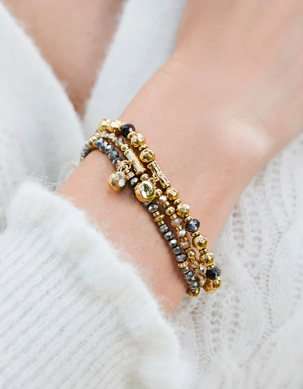 A close-up of a person's wrist wearing a stack of beaded bracelets, including gold, silver, and dark brown beads, styled with a white knit sweater.