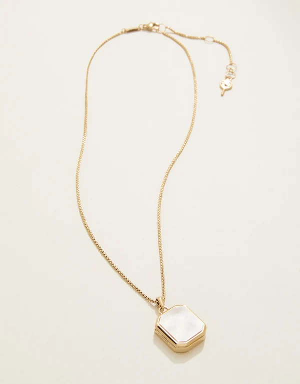 A gold chain necklace with a hexagonal pendant featuring a white mother-of-pearl inlay, displayed on a clean off-white background.