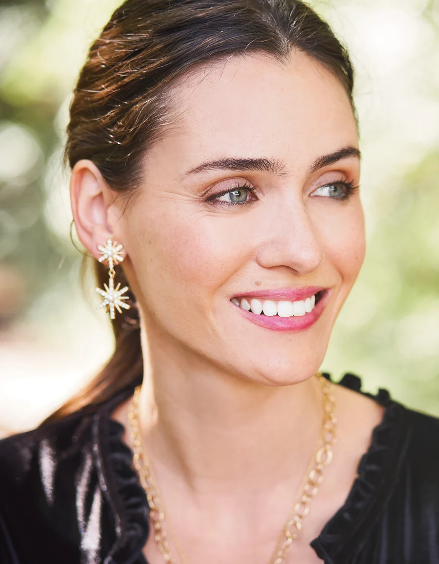 A close-up portrait of a woman with wearing gold starburst dangle earrings, set against a soft, blurred green outdoor background.