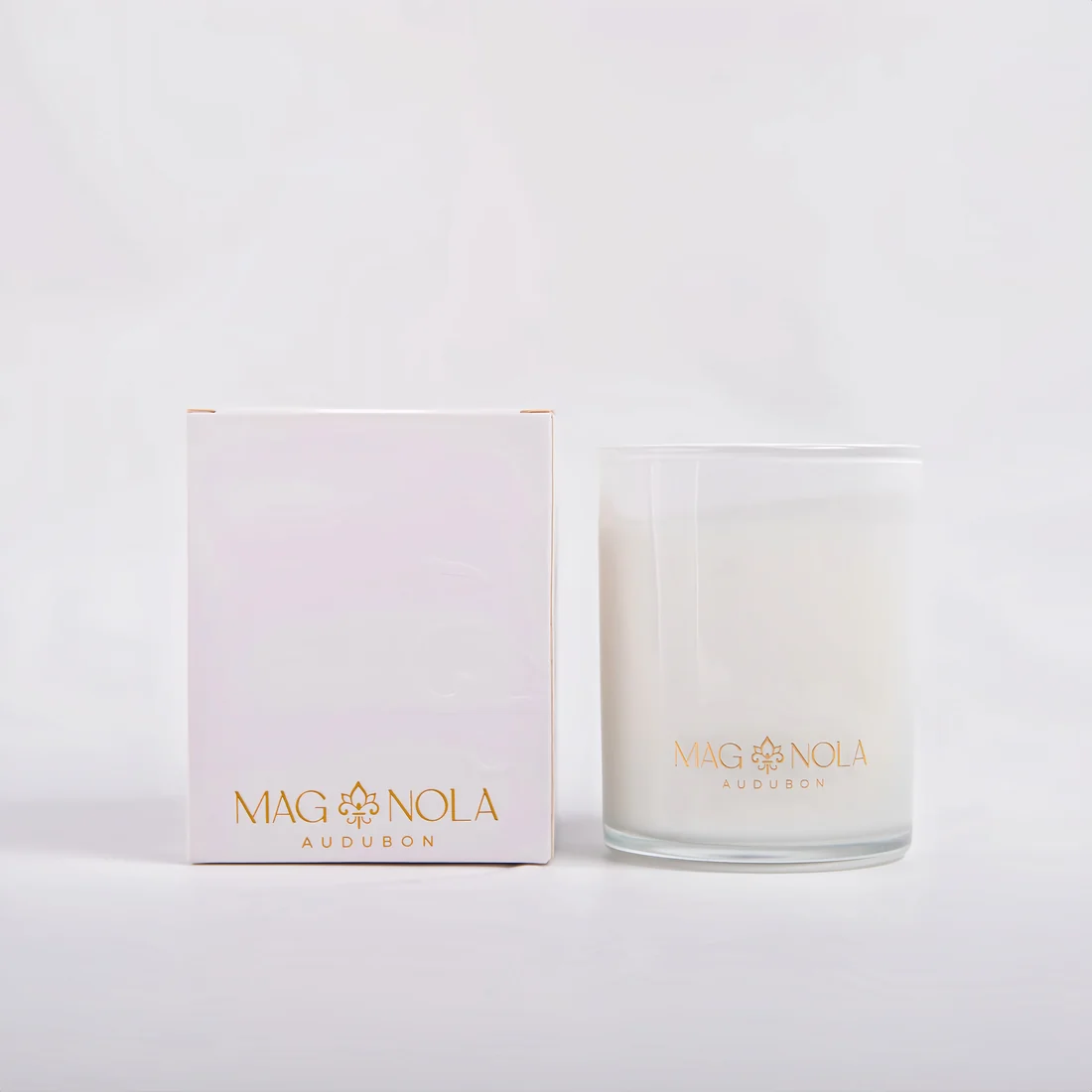 A white 8.5 oz wax candle in a clear glass jar with a gold logo, standing next to its pale pink and white packaging box. The brand name 'MAG NOLA AUDUBON' is printed in gold on both the box and the jar.