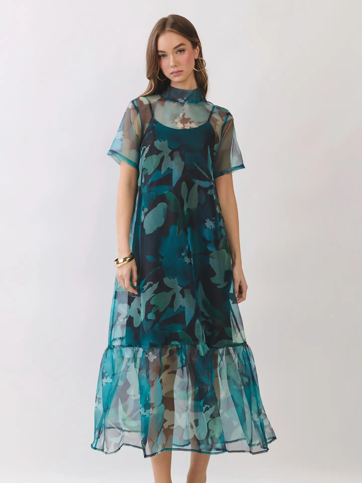A woman modeling a sheer midi dress with a blue and green floral print, featuring short sleeves, a high-neck mesh overlay, and a tiered ruffled hem, set against a plain light gray background.